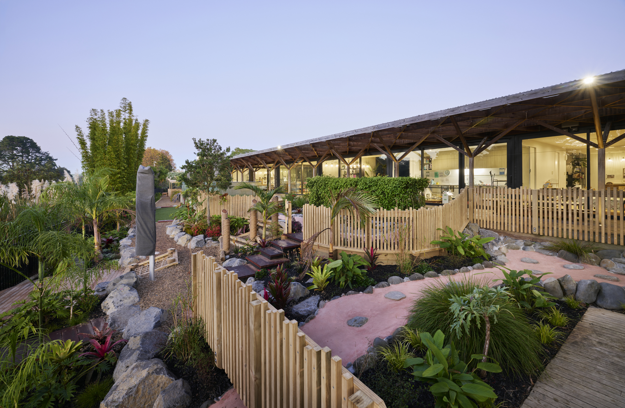 Elevated view of playground with timber fencing, stone boulders, tropical plantings and covered pergola along building