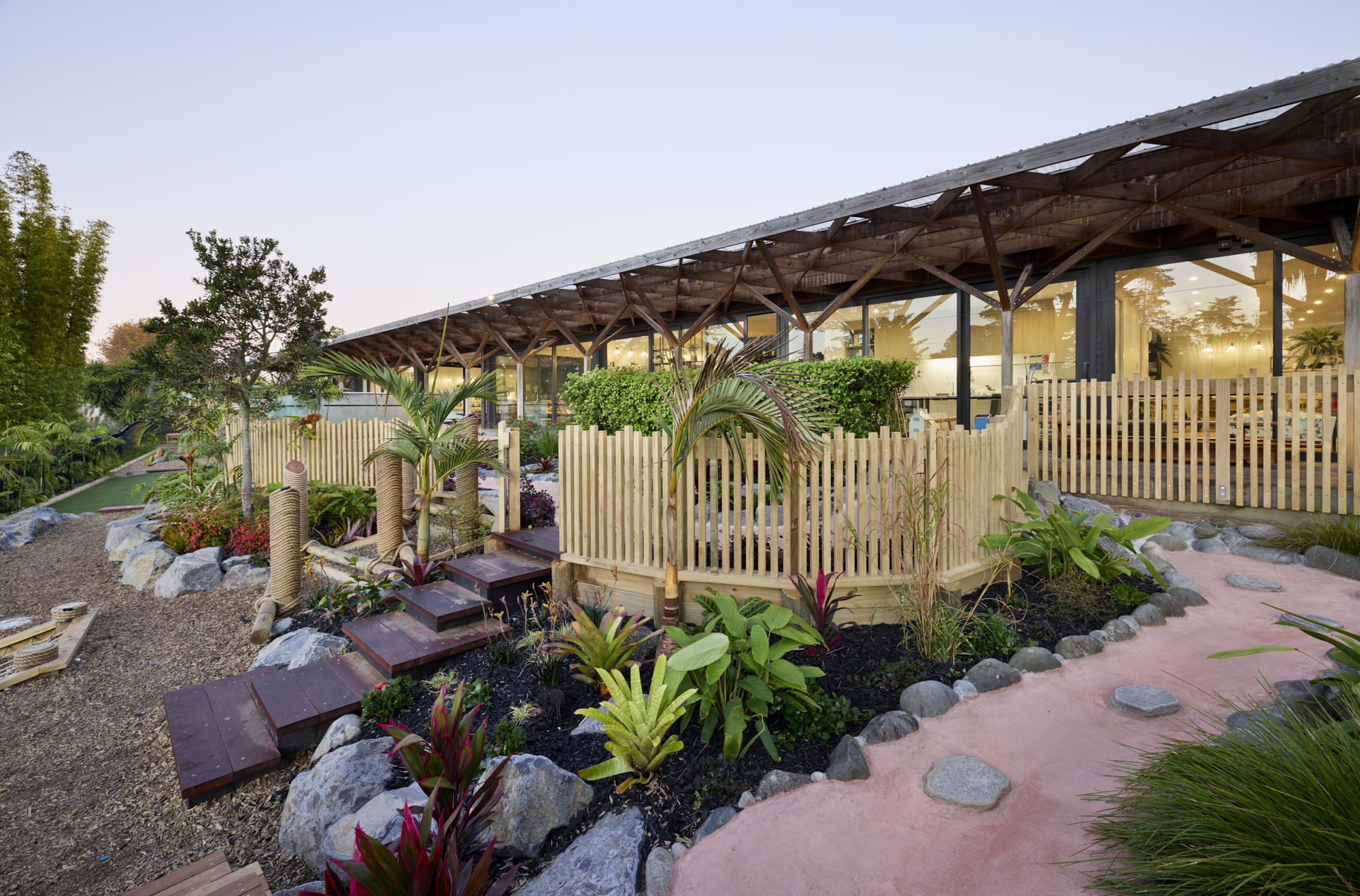 Coloured concrete pathway with stone boulders, young palm trees, red-leafed plants and timber pergola shade structure