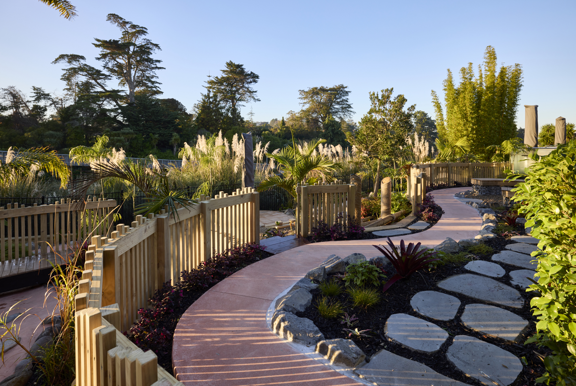 Winding coloured concrete pathway with timber fencing, dark-leafed plantings, stone stepping pads and ornamental grasses