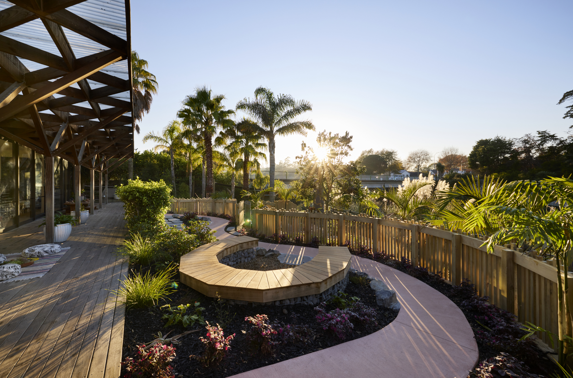Curved timber boardwalk with circular raised seating bench, palm trees and planted garden beds