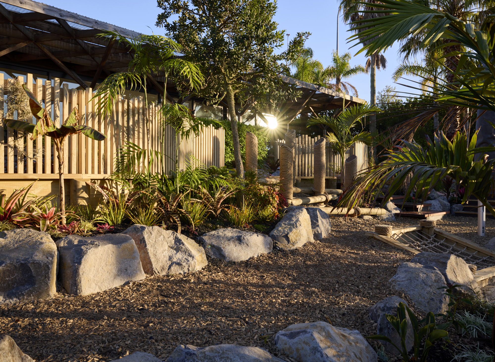 Large stone boulders on bark mulch with tropical plantings, palm trees and timber fencing in afternoon light