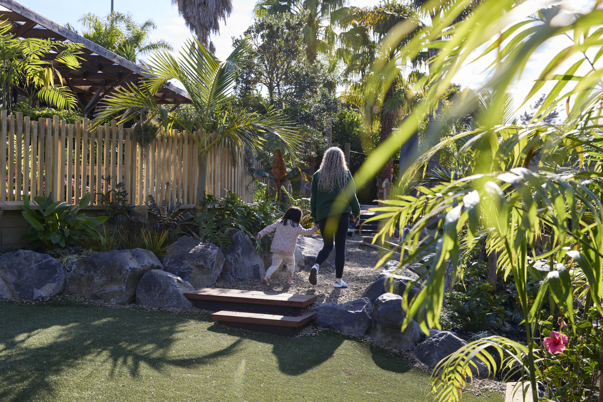 Adult and child walking through playground with stone boulders, bark mulch, palm trees and synthetic turf