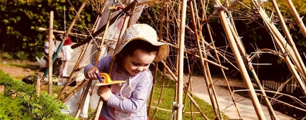 Child in a sun hat using a hand drill beside a bamboo teepee